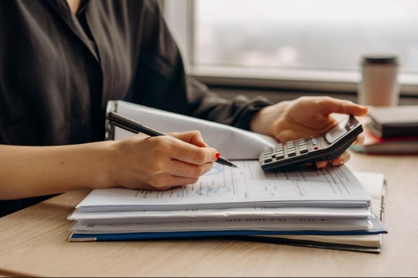 Person holding a calculator in one hand and a pen in another while both hands rest on an open workbook.