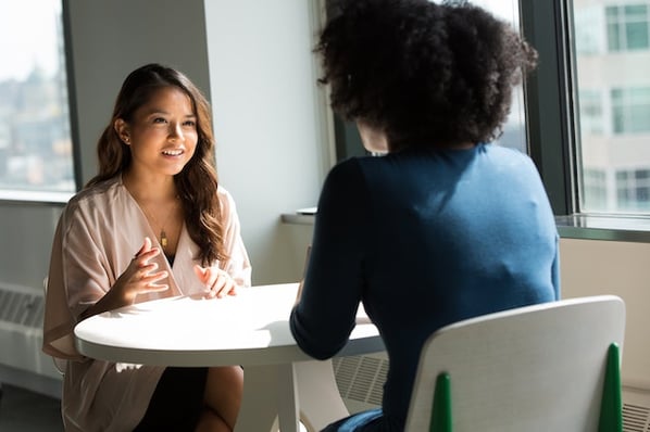 woman in light blouse sits at white table talking to woman in blue shirt