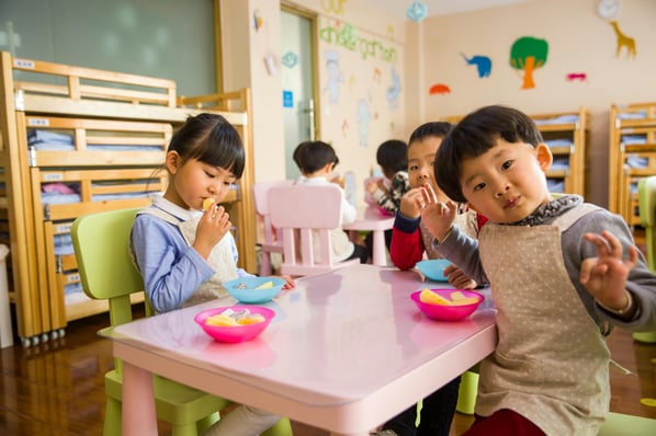 Three toddlers eating lunch at a small pink table in a preschool classroom.