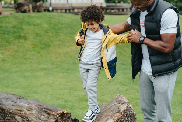 A father helps his young son balance as he walks on top of a large, fallen tree trunk in a park.