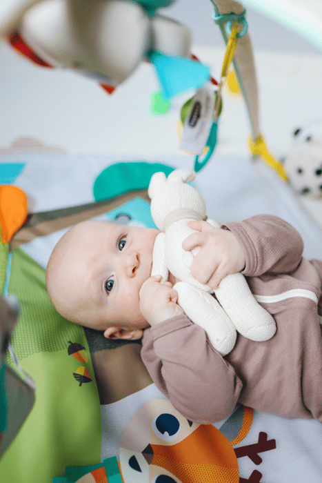 An infant wearing a brown onesie and laying on a blue and green play mat. He is holding a white toy rabbit. The rabbit's arm is in his mouth.